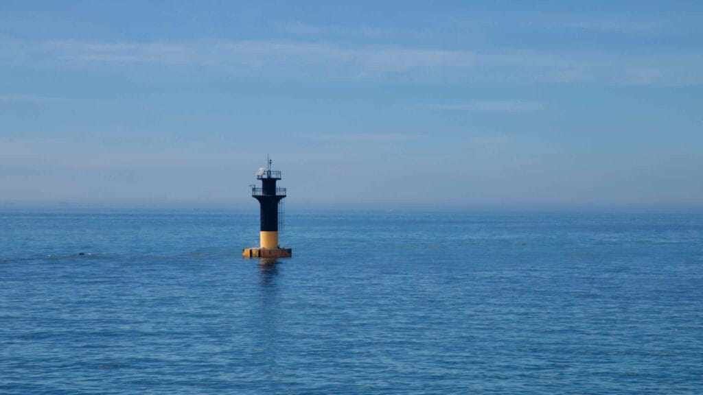 A black‑and‑yellow offshore beacon rises from the Korea Strait near Iho.