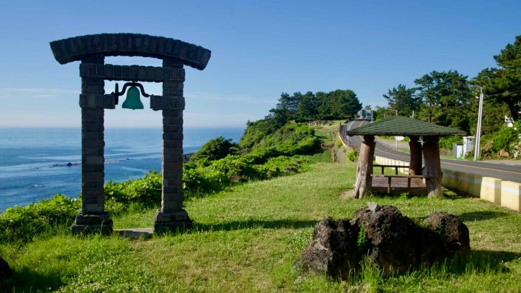 A small clifftop park on the Hagu‑Aewol Coastal Road features a bell set in a basalt arch and a rustic wooden shelter.