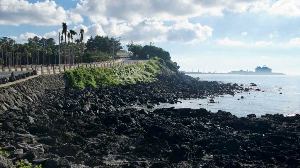 A winding section of the Hagu‑Aewol Coastal Road follows a cliff above dark basalt rocks.