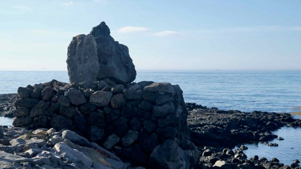 Basalt rocks frame a weathered stone monument on the shoreline near Gonae Port.