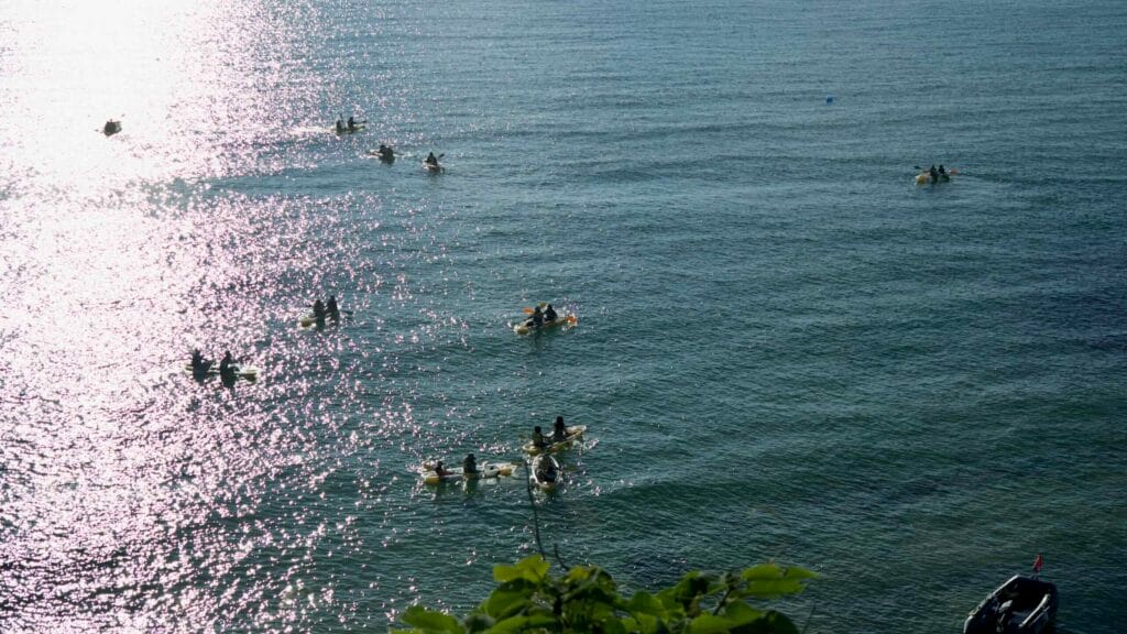 Groups of kayakers paddle across sun‑sparkled water near Handam Beach.