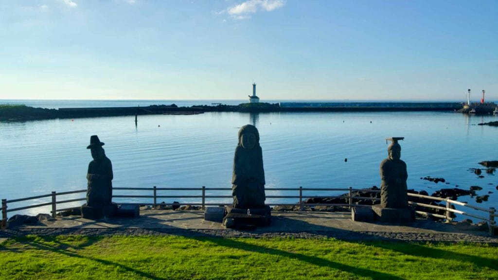 Stone figures at Yeongdeung Halmang Myth Park facing a calm harbor and lighthouse.