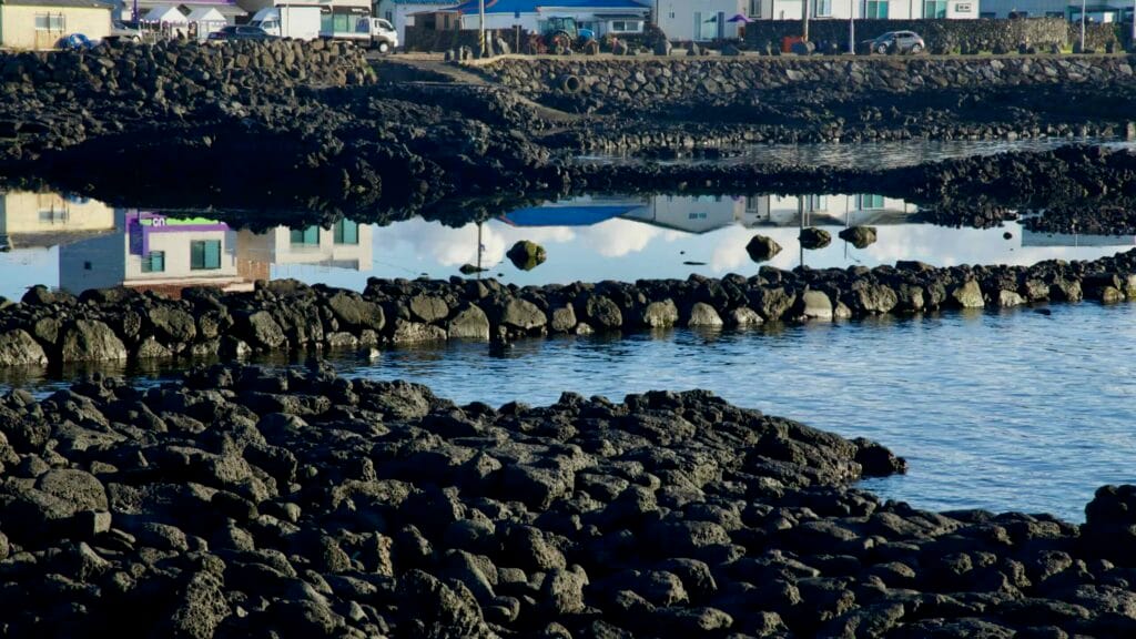 Basalt fish weirs reflect village buildings along the inlet at Gwideok.