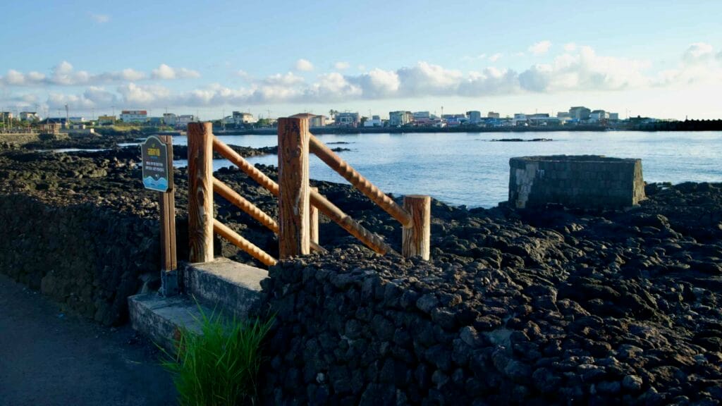 Wooden steps over a lava‑stone wall lead down to the rocky shore at Gwideok.