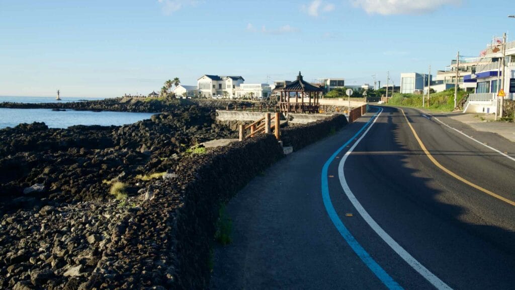 A bike lane and two‑lane road follow a basalt seawall past a wooden pavilion.