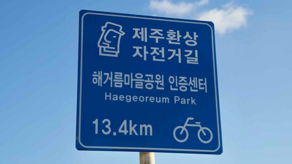 Blue road sign for the Jeju Fantasy Bike Path indicates 13.4 km to Haegeoreum Park.