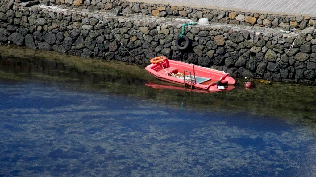 A small red dinghy is tied beside a basalt quay in clear tidal water.