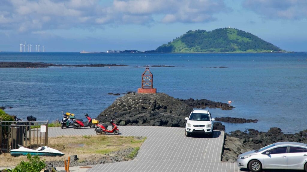 Scooters and cars park on a lava-rock pier leading to a small steel beacon.