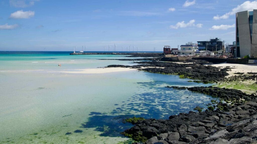Hyeopjae shoreline with lava rock, pale sand and distant windmills.