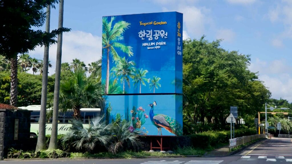 Roadside entrance sign for Hallim Park featuring a peacock mural beside palm trees.