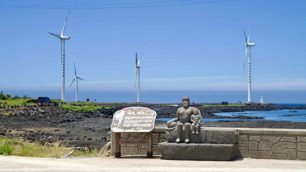 Haenyeo statue and sign at Sinchang with wind turbines across lava flats.