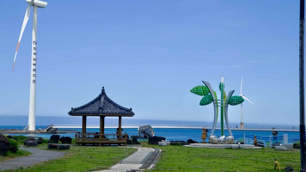 Gazebo and green sculpture by the sea with turbines and bridge.