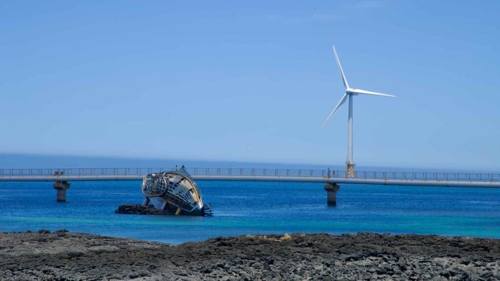 Metal shipwreck sculpture with offshore turbine and pedestrian bridge.