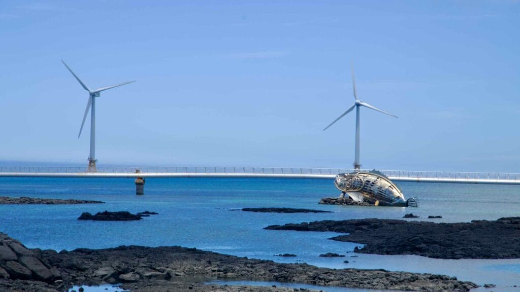 Leaning shipwreck beside the ocean walkway with twin turbines beyond.