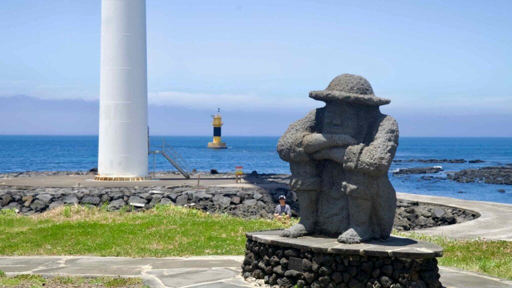 Dolhareubang statue by the sea near a turbine base and beacon.