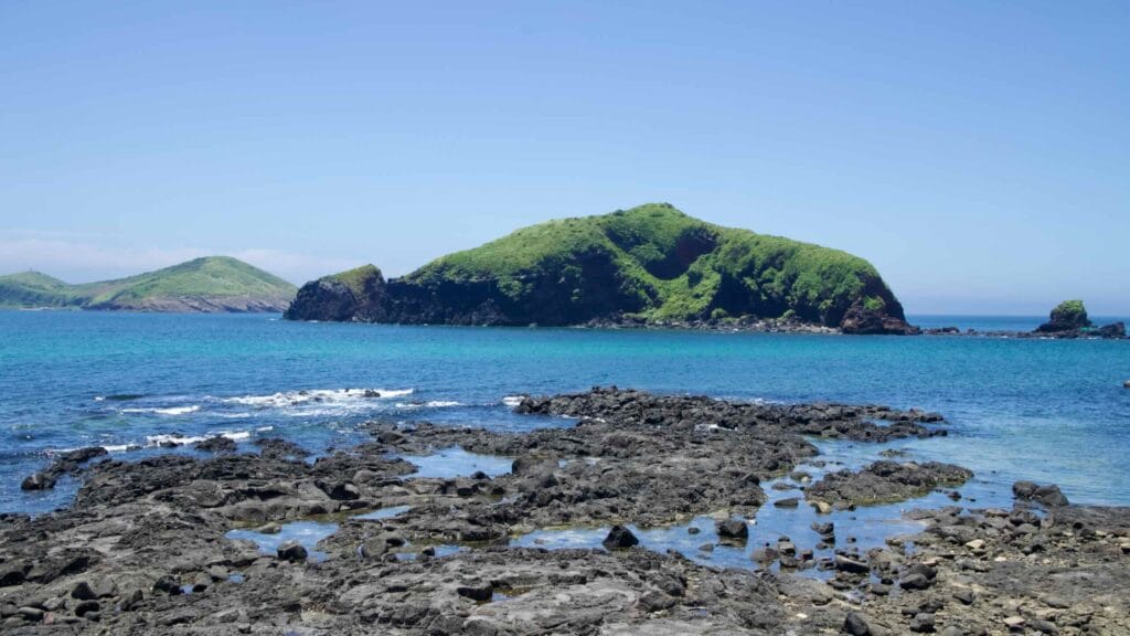 Basalt tide pools and a green islet off Chagwido under clear summer skies.