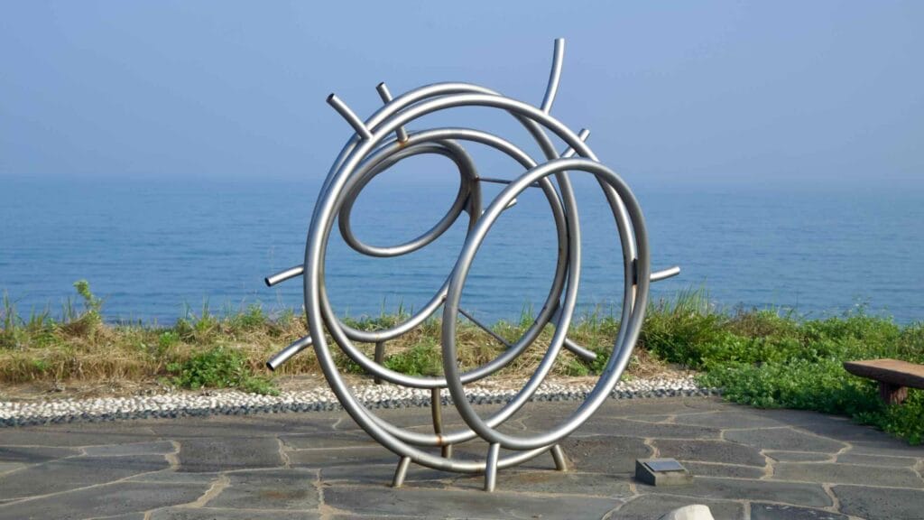 Abstract stainless‑steel sculpture beside the seaside promenade on Brother’s Coast Road.