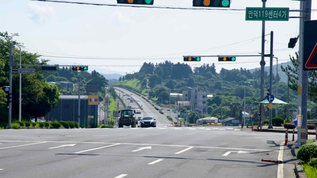 Traffic pauses at a broad Iljuseo Road intersection in Andeok-eup, dropping toward forested hills.