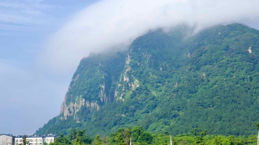 A cap cloud drapes Sanbang Mountain’s cliffs above apartments and pine forest on Jeju’s south coast.