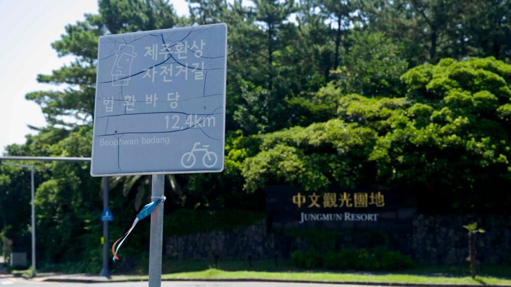 A weathered Jeju cycling route sign points toward Beophwan Badang near the Jungmun Resort stone wall.