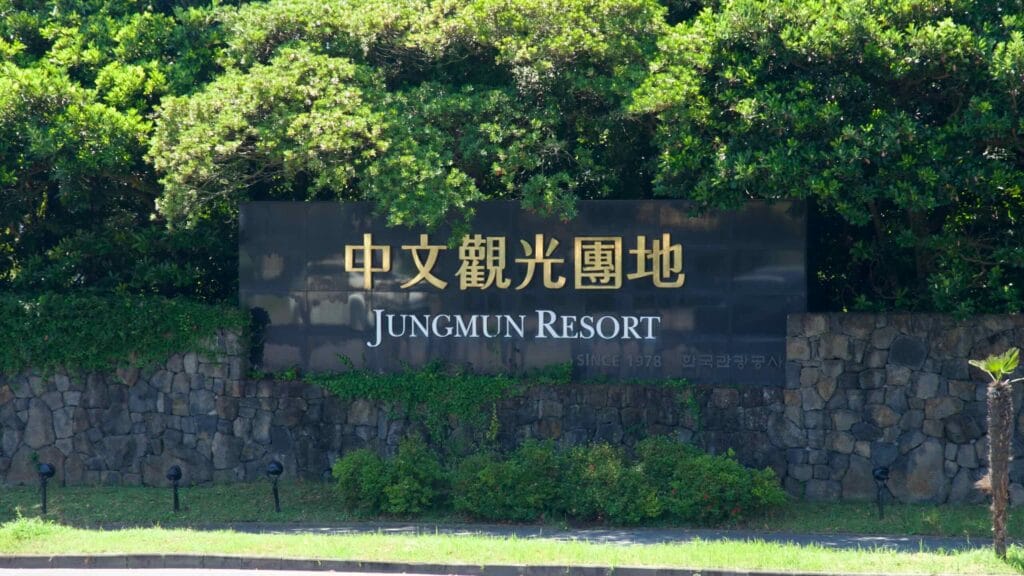 The entrance sign for Jungmun Resort stands on a black panel set into a basalt wall, surrounded by palms.