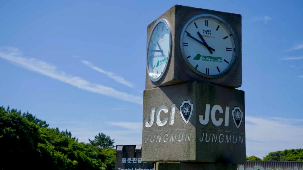 A cube clock marked JCI Jungmun stands above a tourist plaza, with twin dials and nearby branding.