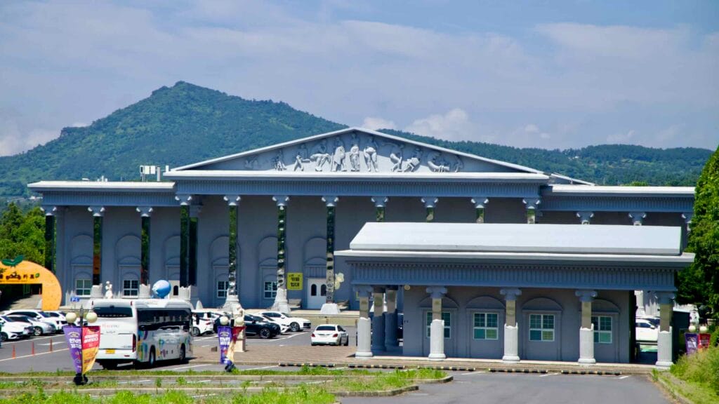 A museum building with columns and a pediment relief stands before parking buses and visitors’ cars.