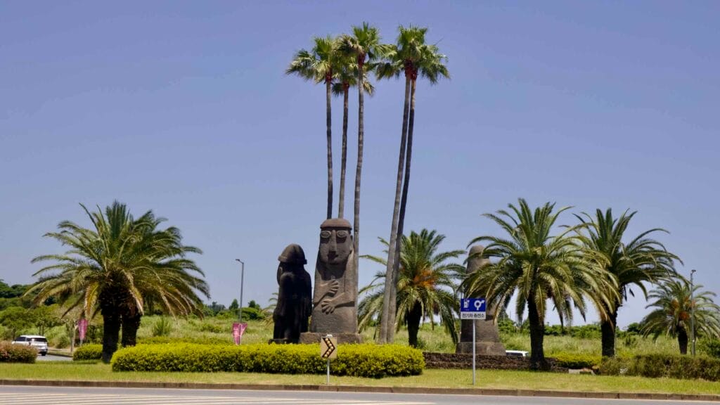 Dol hareubang statues rise among palms at a landscaped Jungmun roundabout.