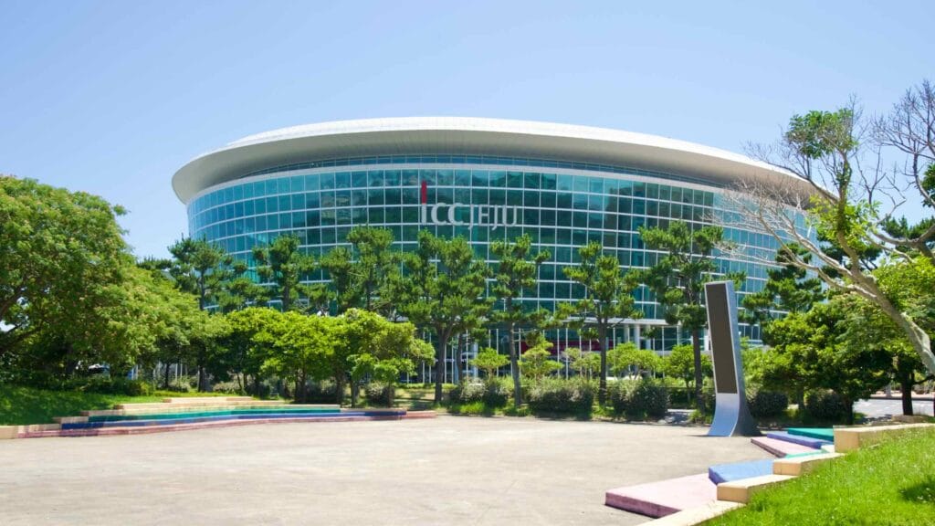 ICC Jeju’s curved glass facade fronts a wide plaza with steps and pines.