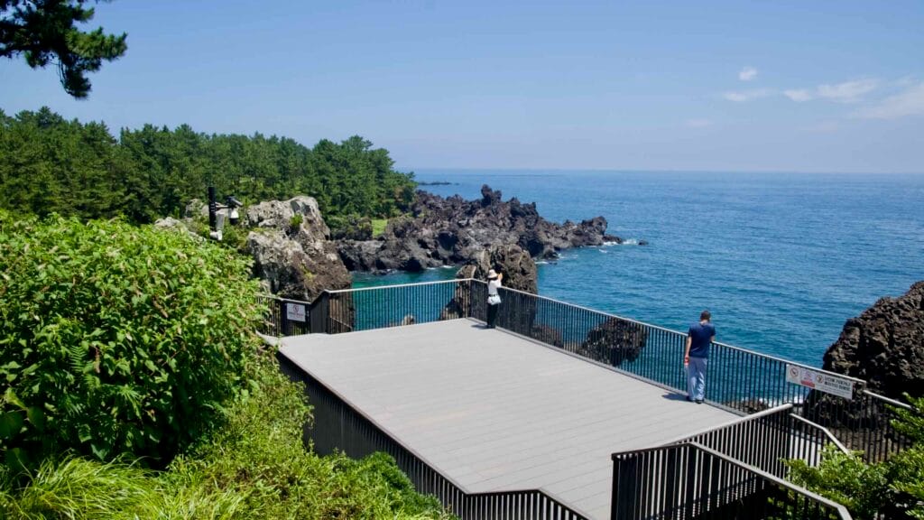 Visitors stand on a deck above the Daepo Jusangjeolli basalt cliffs and turquoise waters.