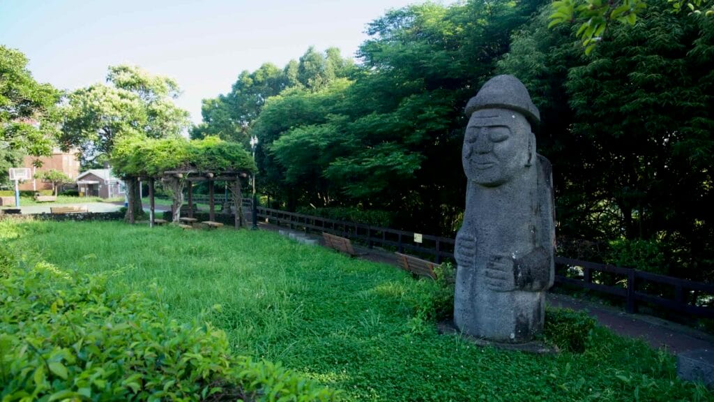 A basalt dol hareubang stands by a shaded lawn and walkway in Chilsimni Park.