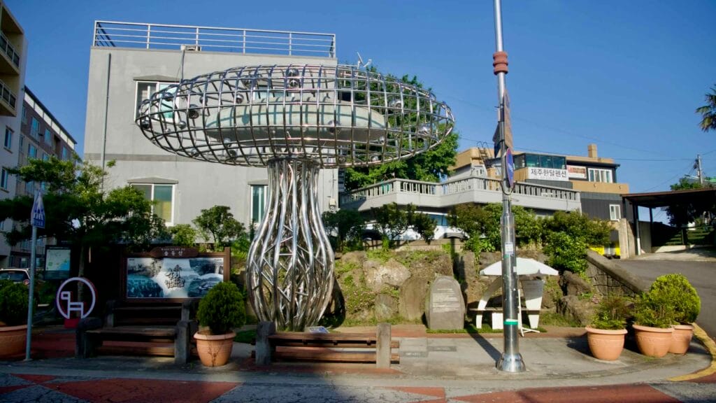 A dome‑like steel sculpture anchors a small plaza with benches and nearby shops in central Seogwipo.