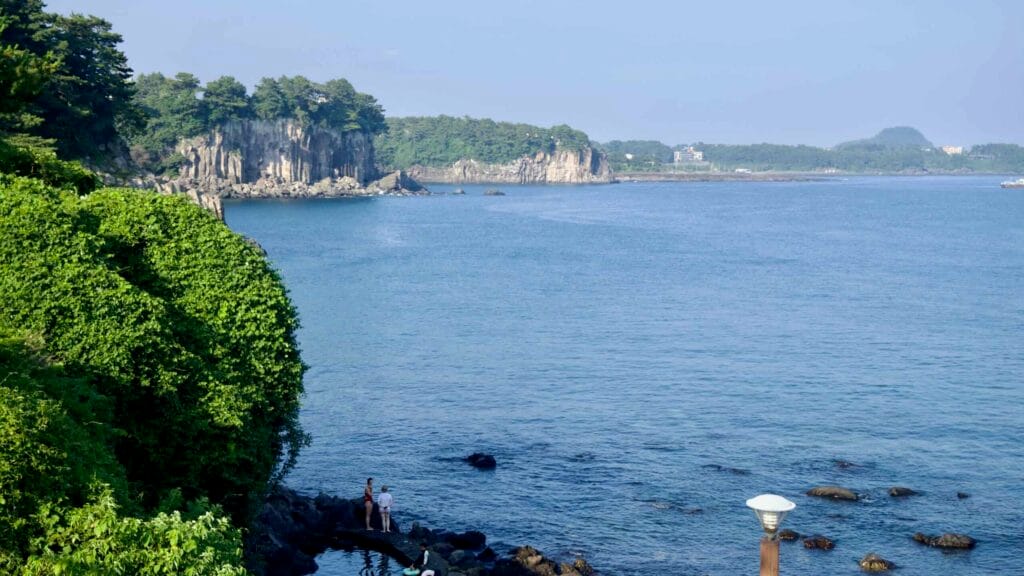 Basalt headlands and calm inlets along the Seogwipo coast with visitors on tidal rocks.