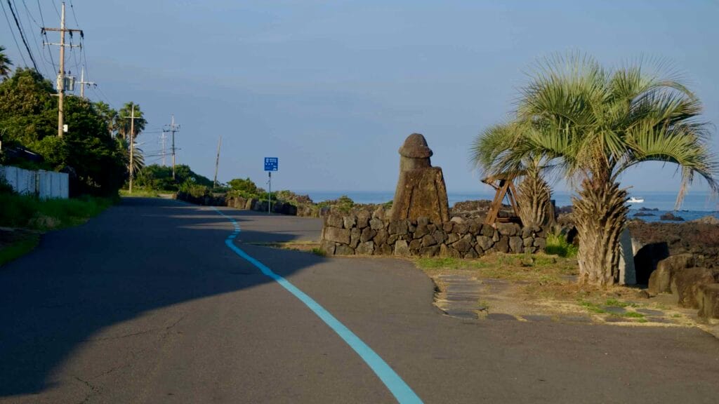 A dolharubang stands beside a coastal road and palms.