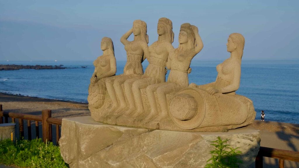 A stone sculpture faces the sea beside Soesokkak’s black‑sand shore.