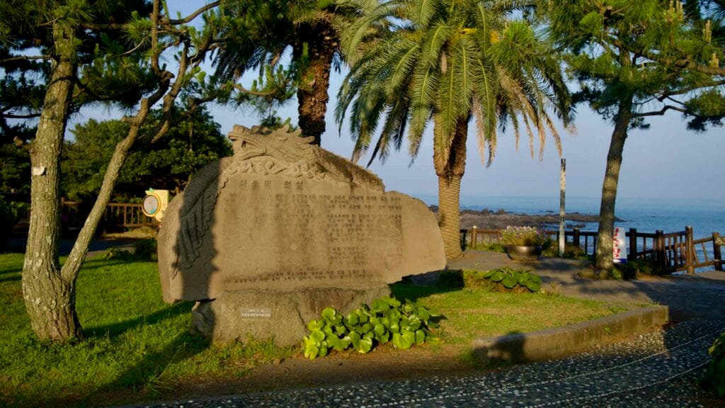 A carved stone monument stands among palms and pines at Soesokkak Park by the sea.