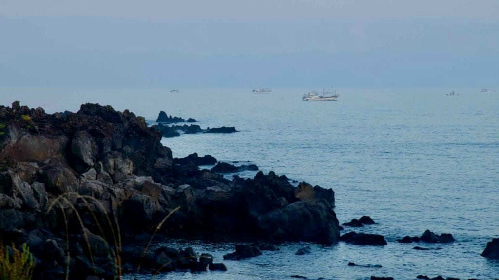 Fishing boats cross calm water beyond the basalt headland near Mangjang Port.