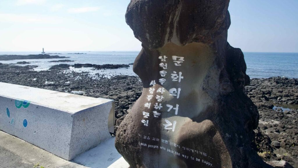 A carved lava‑rock marker faces reefs and a lighthouse on the Namwon coast.