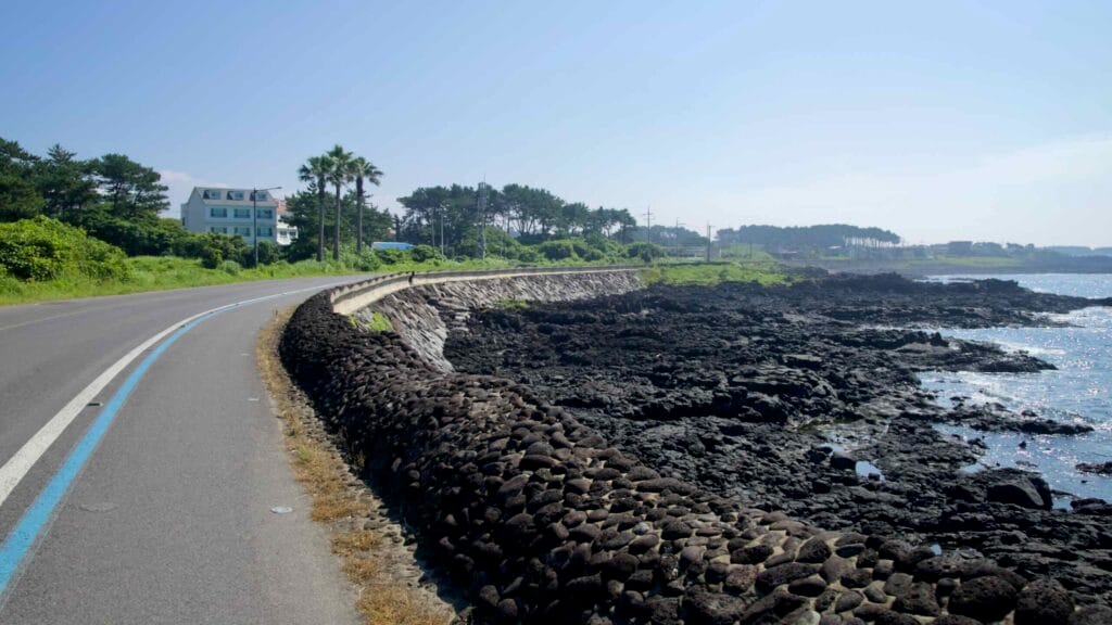 A coastal road curves by a basalt seawall with lava rocks and calm surf near Taeheung.