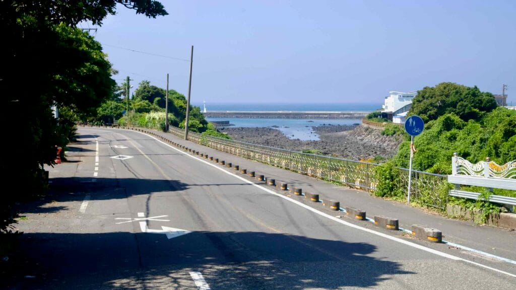 A two-lane road descends toward a rocky inlet near Sinheung-ri, with a bike sign and breakwater beyond.
