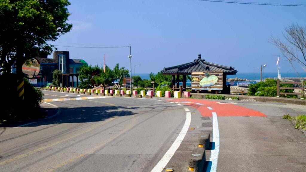 A curve in the road passes an information pavilion above Sinheung-ri Port with bollards.