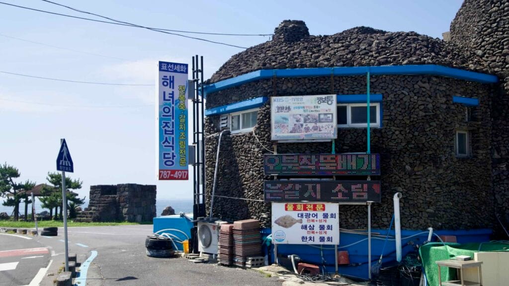 A stone-clad restaurant advertising haenyeo seafood stands by the road at Sinheung-ri, with signs and sea beyond.