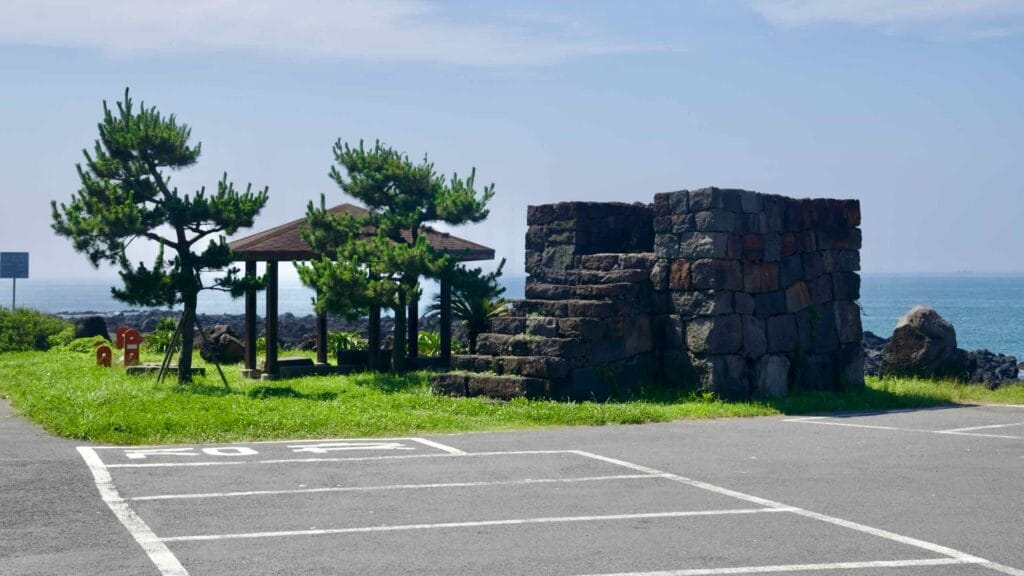 A square stone platform and shade pavilion stand by wind-shaped pines on the basalt shore near Sinheung-ri.