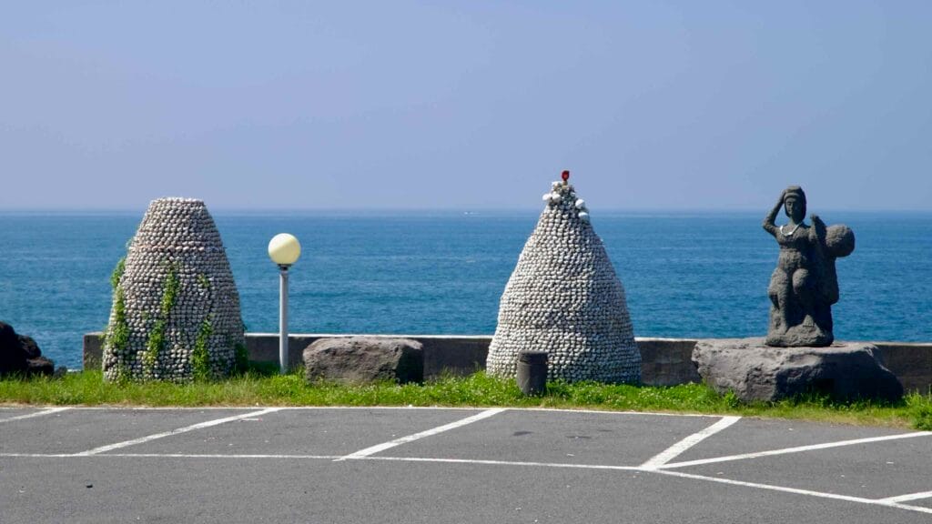 Two shell-covered cairns and a haenyeo diver statue face the sea on a turnout near Sinheung-ri with parking ahead.