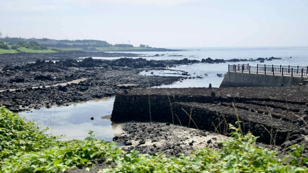 Shallow tidal pools and stone walls sit on a basalt shore near Sinheung-ri with a small pier reaching to the sea.