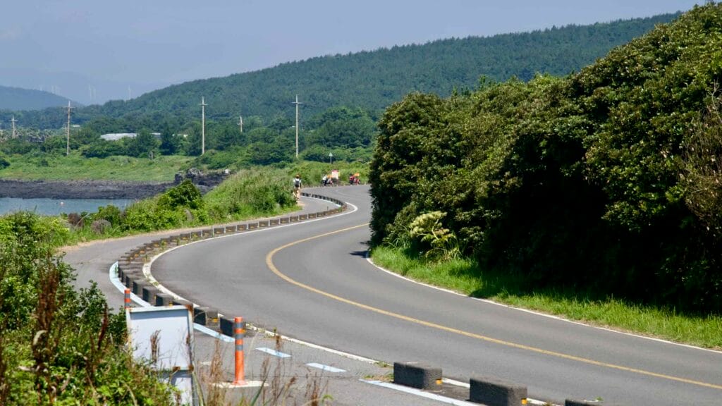 Cyclists ride along a curving coastal road near Sinheung-ri with greenery, barriers, and blue bike markings.