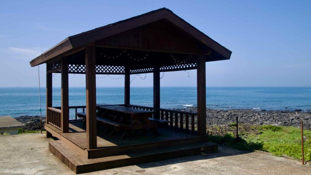 A wooden pavilion stands above the basalt coast near Sinheung-ri, facing calm blue water and low surf.