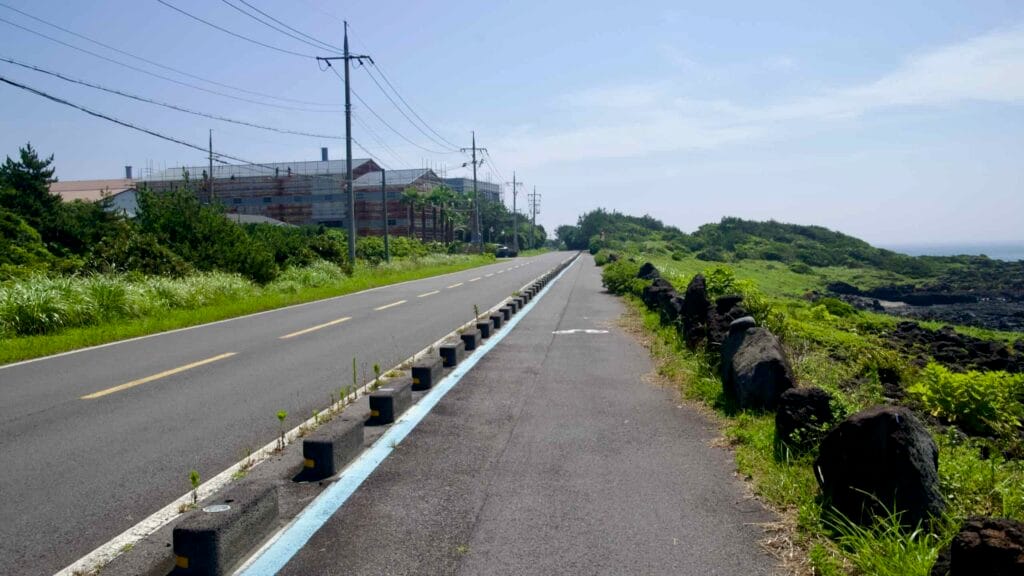 A separated bike lane runs beside a two-lane road on the basalt shore near Sinheung-ri with scaffolding nearby.