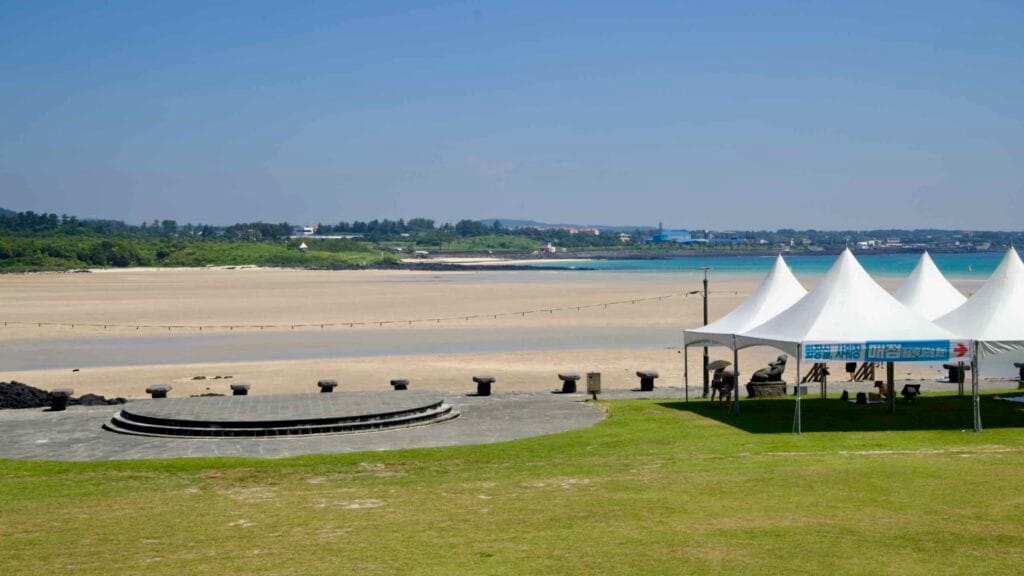 A broad tidal flat stretches across Pyoseon Beach at low tide with an amphitheater and white tents by the sea.