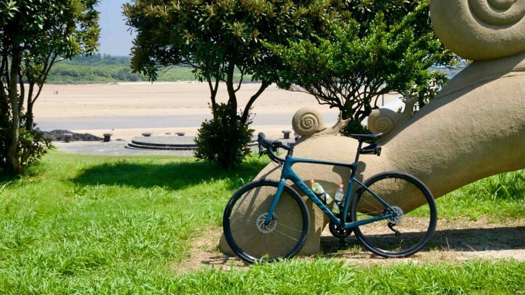 A road bike leans against a snail sculpture in the park above Pyoseon Beach with views of the stage and sand.